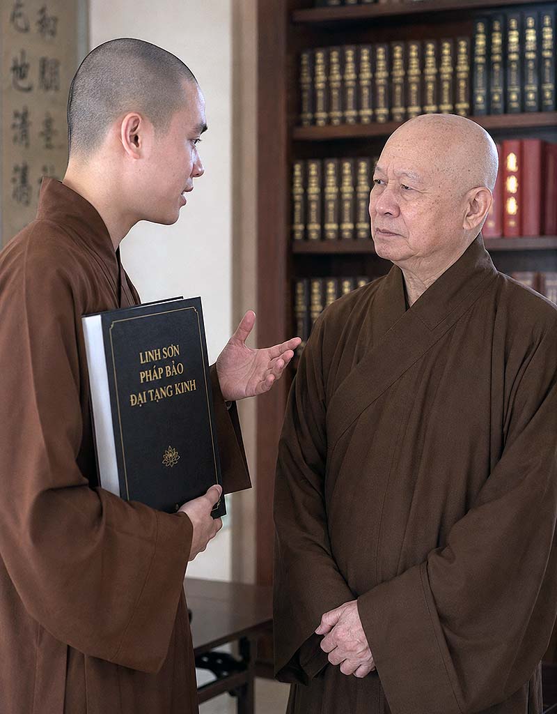 兩位佛教師徒討論佛法，年輕比丘手裡捧著一本靈山法寶大藏經